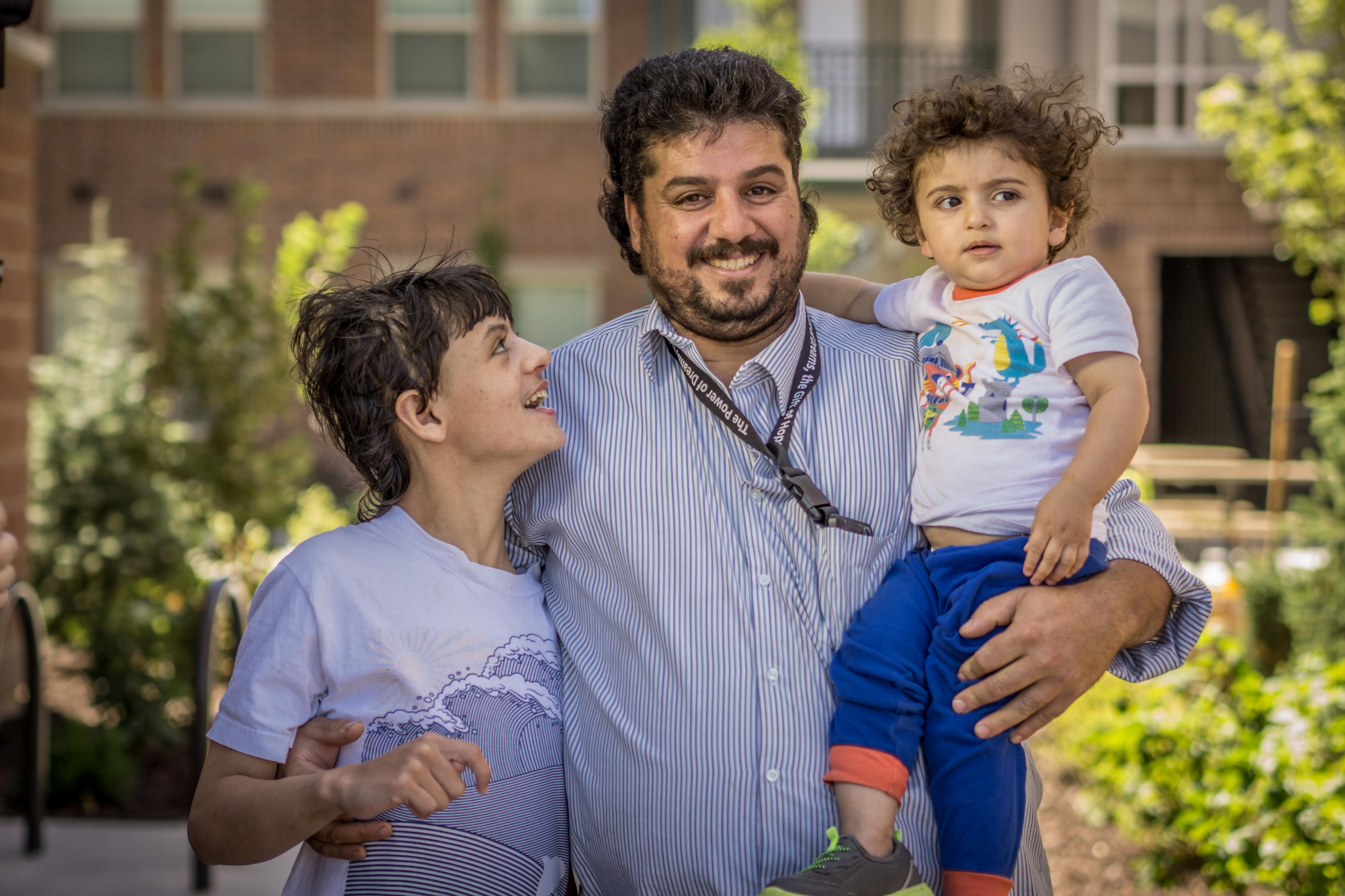 Photo of a father holding his toddler with arm wrapped around older child. Big smiles and a feeling of pride.